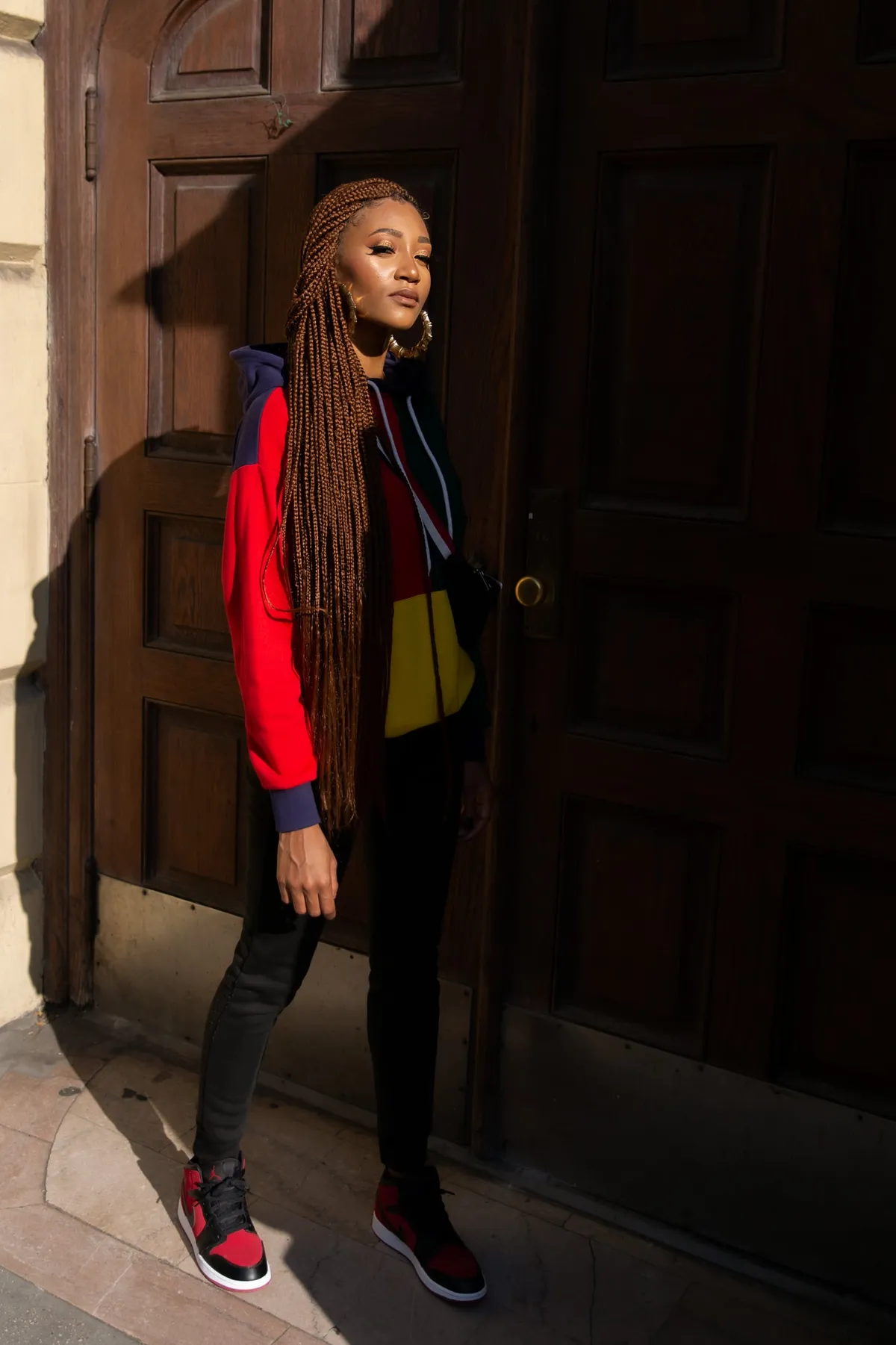 Full-length portrait of a woman in a red, navy and yellow hoodie and red sneakers posing against a dark wooden door