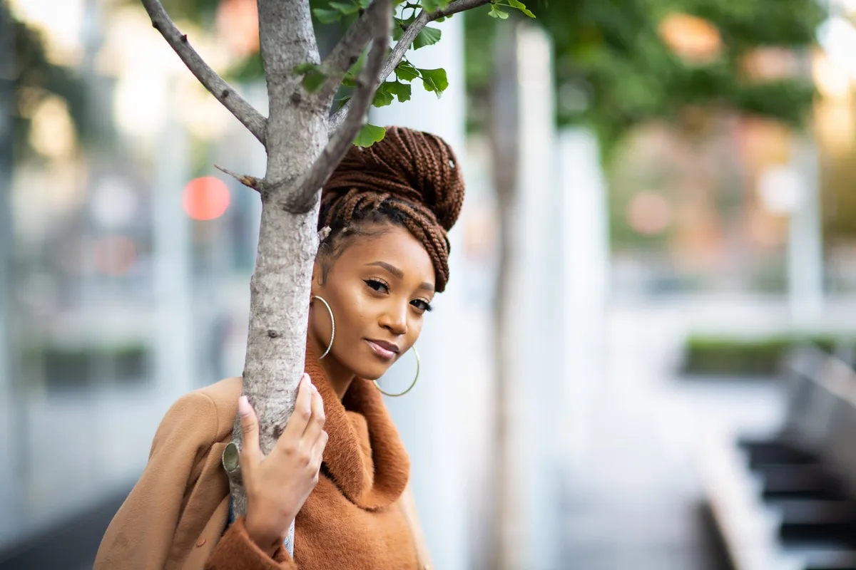 A woman with braids in a high bun peeks around a slim tree trunk on a city plaza