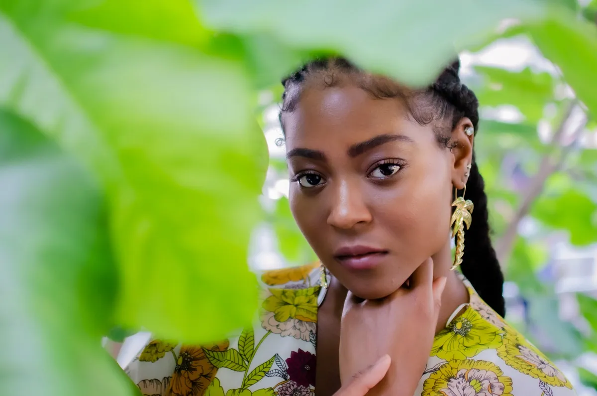 A woman in a floral top looks through large green leaves, her hand at her neck