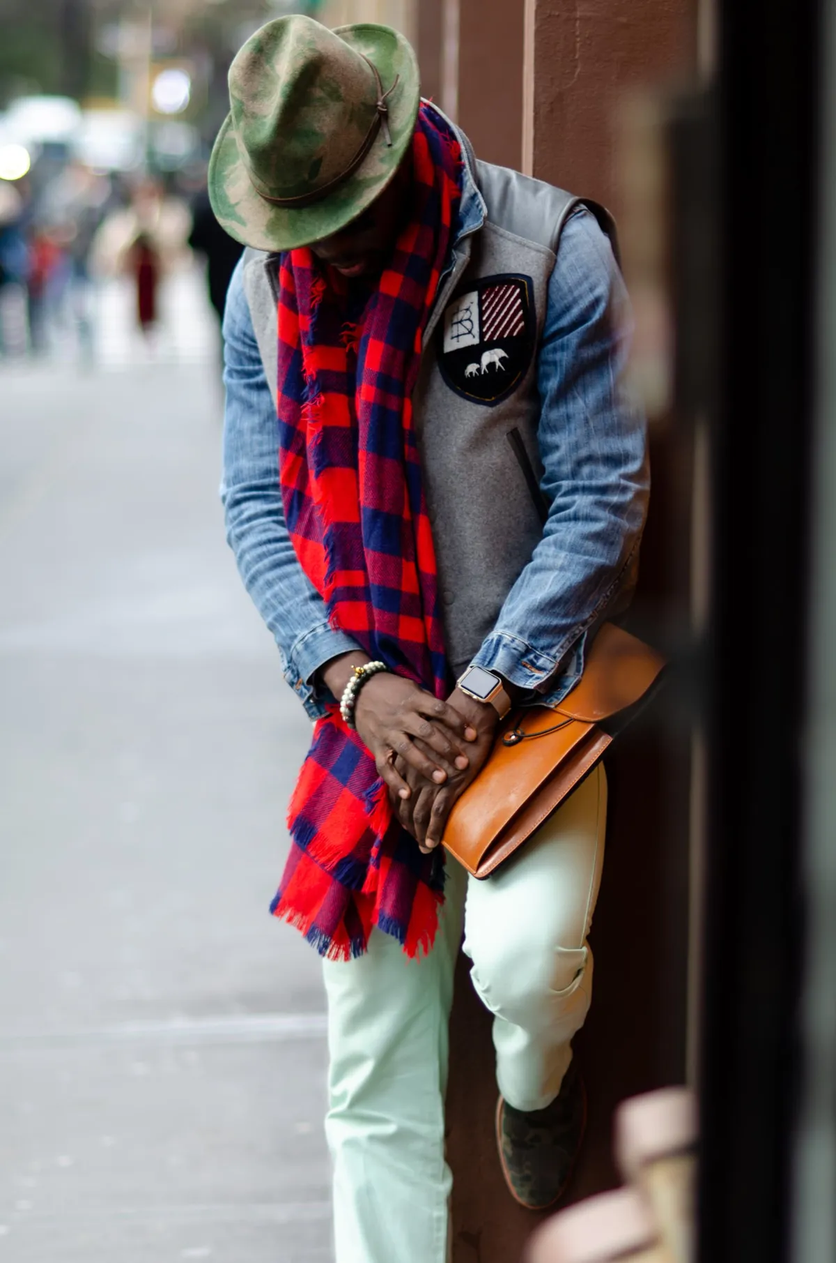 A man in a fedora and plaid scarf looks down while leaning against a storefront, a leather bag at his side