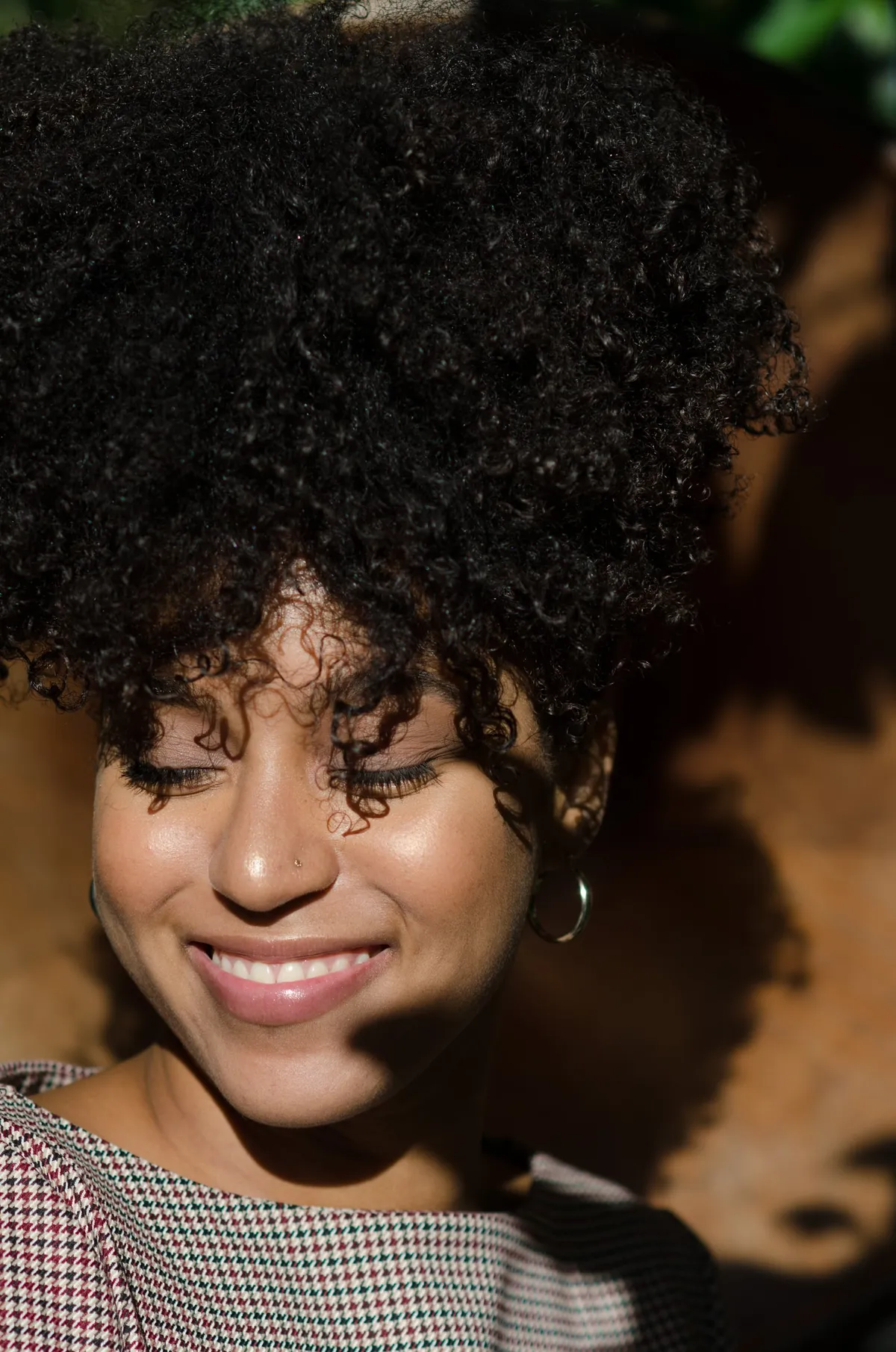 A woman with curly hair smiles with her eyes closed in warm dappled light