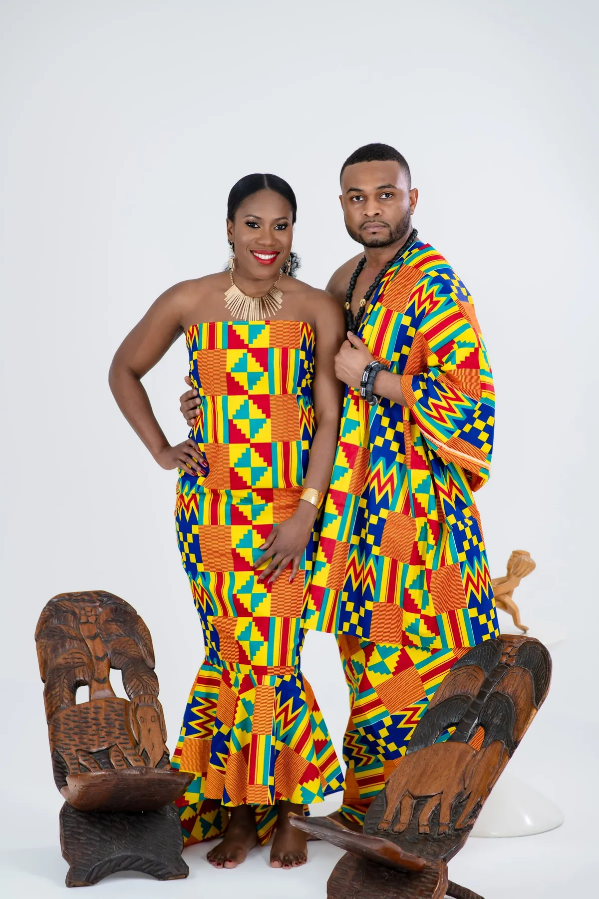 A smiling couple in vibrant kente garments stand together beside carved Ghanaian stools