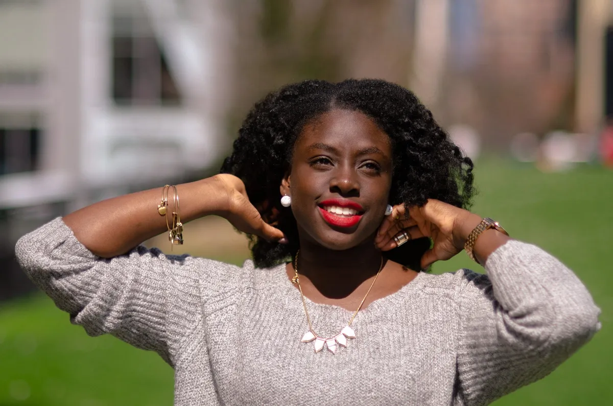 A woman with red lipstick smiles with her hands lifted behind her head in bright sun