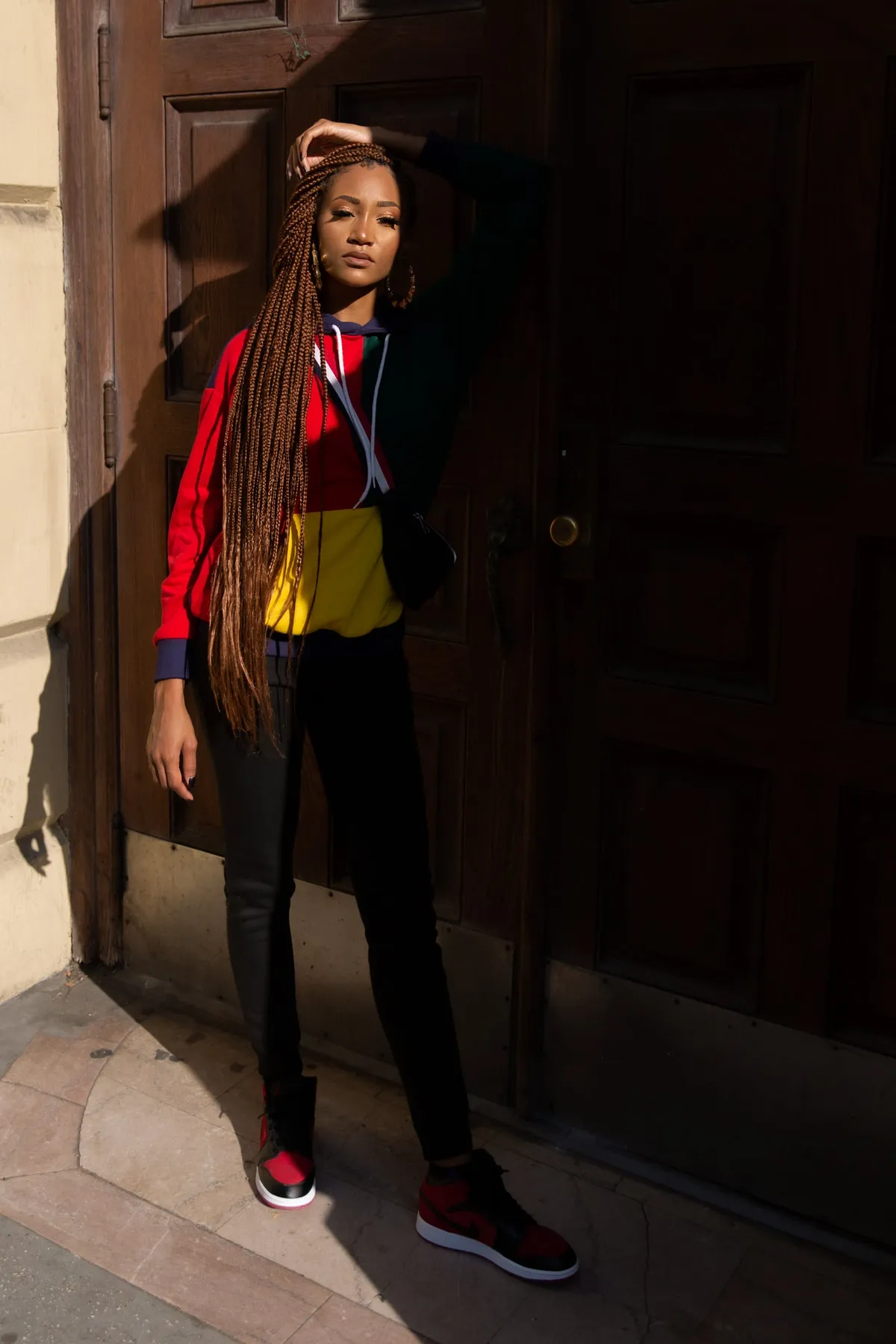 A woman with waist-length braids touches her hair while standing in a sunlit doorway in black jeans and red sneakers