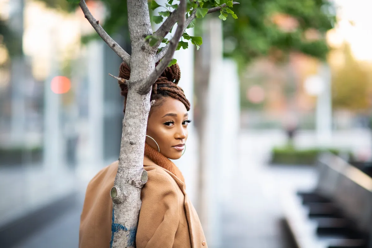 A woman in a camel coat leans against a tree trunk with a blurred city plaza behind her