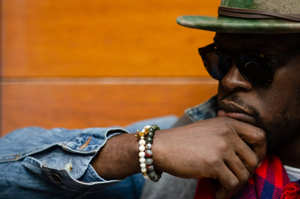 Close-up of a man in sunglasses and a denim jacket resting his chin on his hand, a beaded bracelet on his wrist