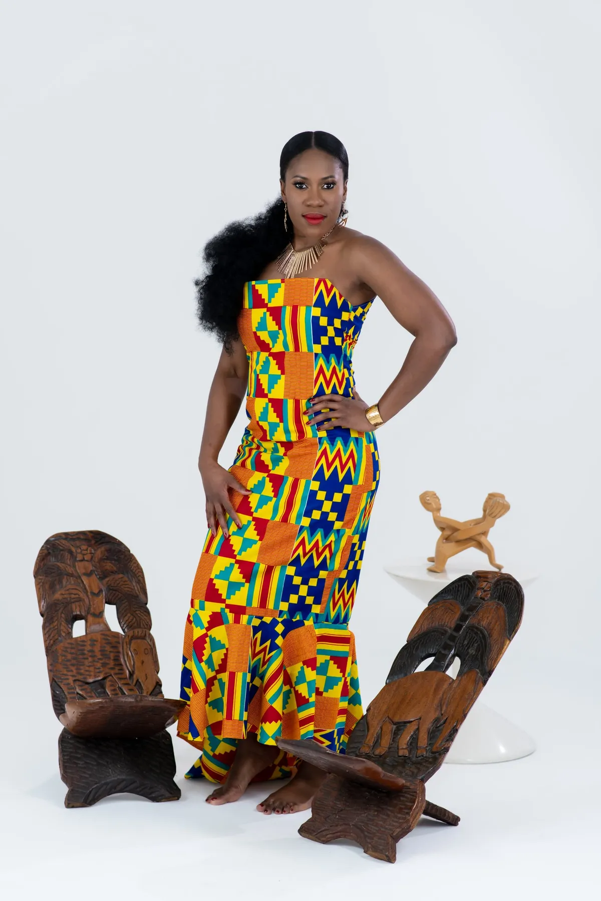 A woman in a kente mermaid gown poses with a hand on her hip between carved wooden chairs