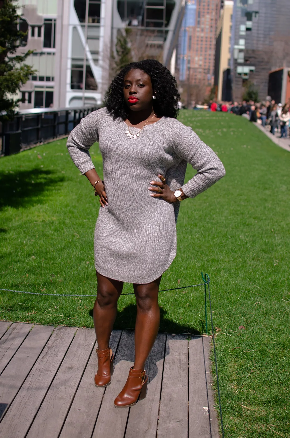 A woman in a grey sweater dress and brown boots stands with hands on her hips on the High Line