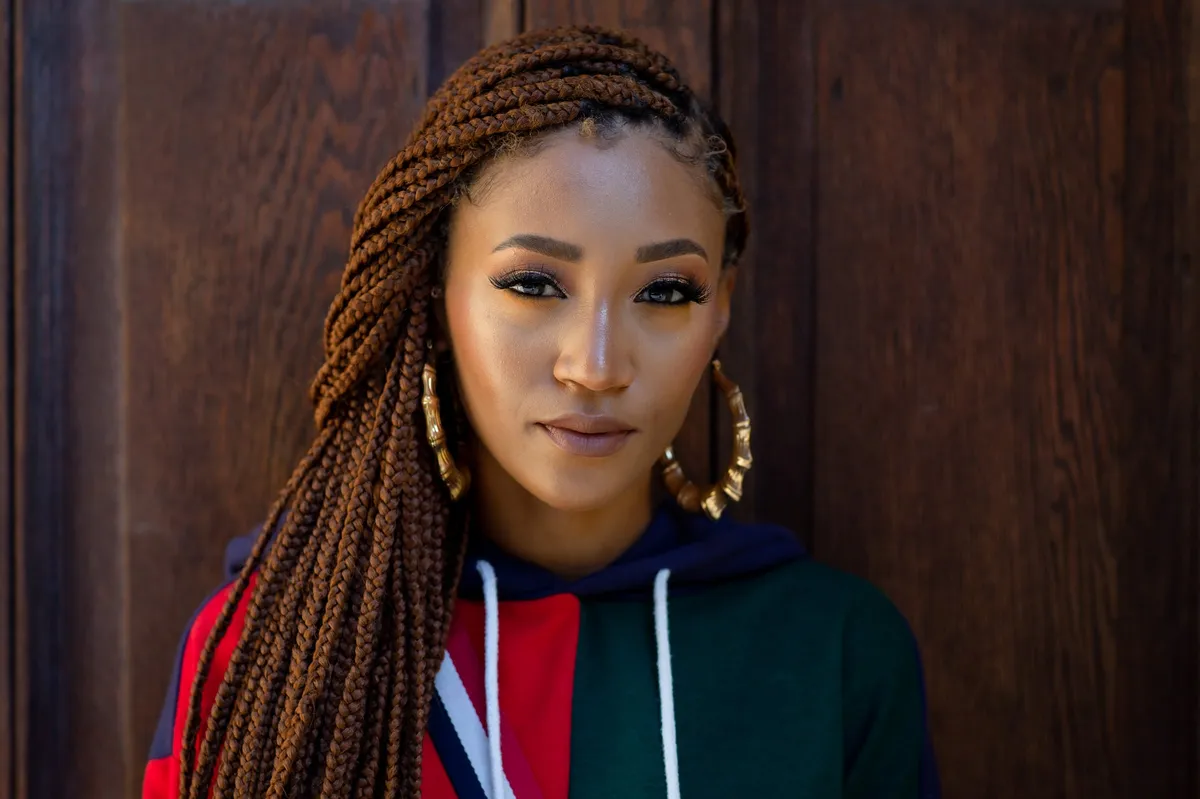 Close-up of a woman with auburn braids and gold hoop earrings gazing into the camera against warm wood