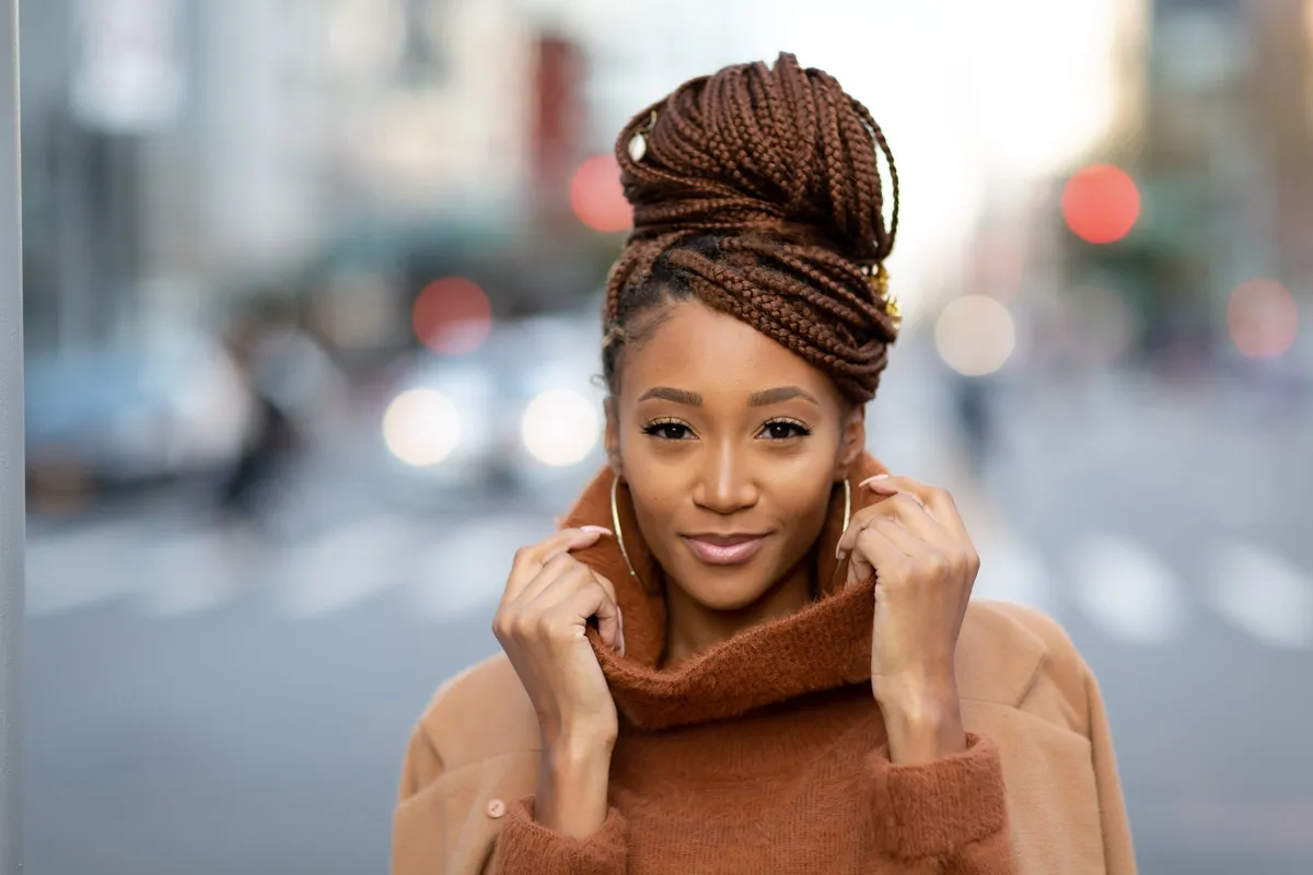 A woman holds the collar of her rust turtleneck, the city street softly blurred behind her