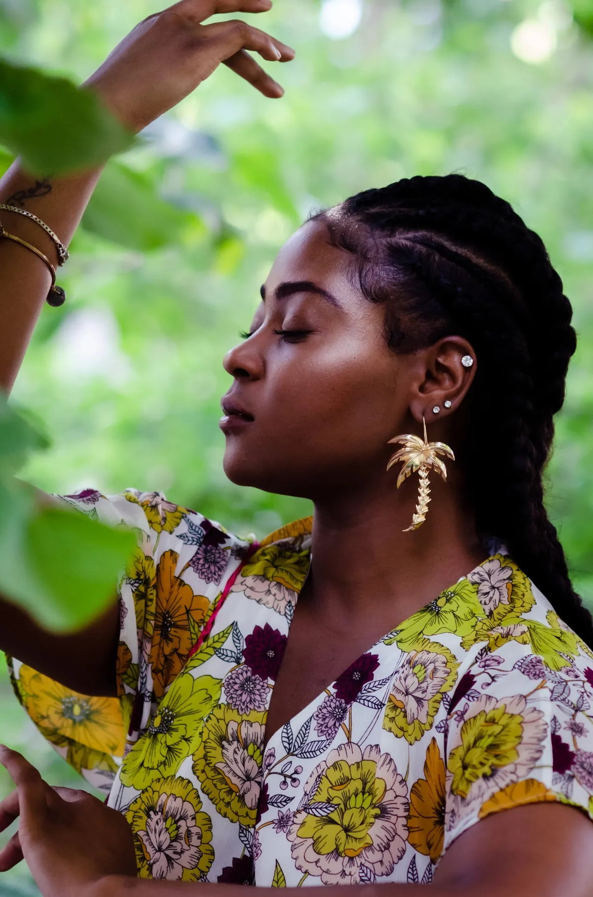 A woman in a floral blouse raises her arm among green leaves, gold earrings in profile