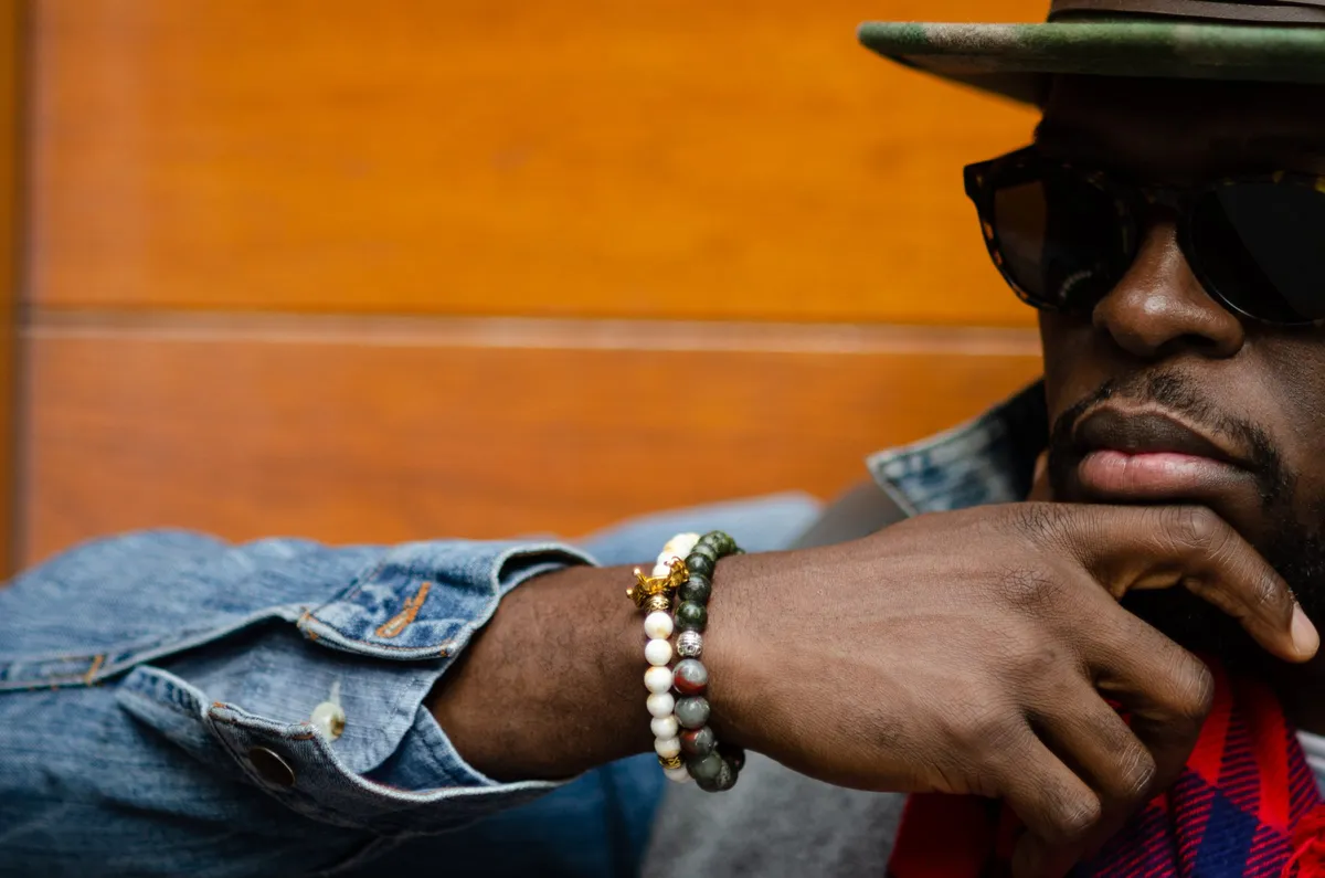 A man in dark sunglasses and a fedora rests his chin on his hand against warm orange wood paneling