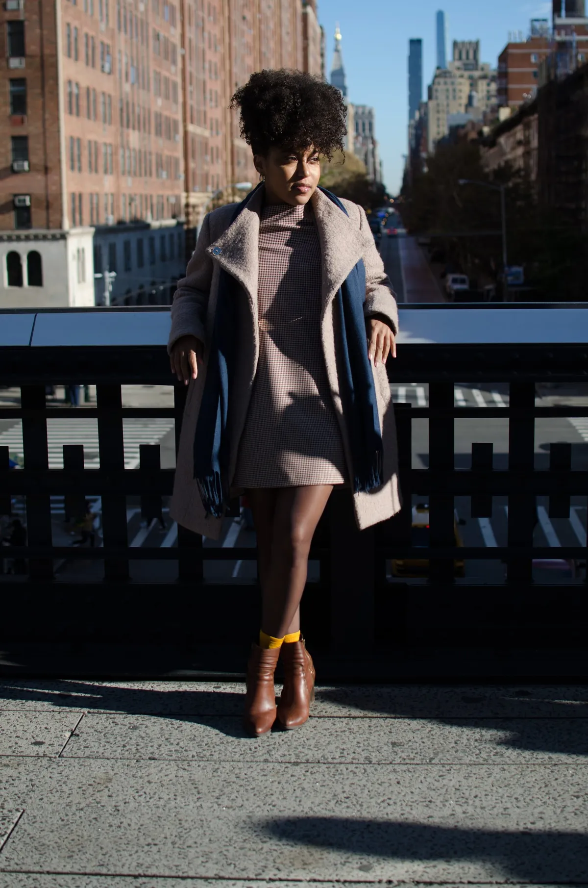 A woman in a long coat stands on a High Line overlook with the city behind her
