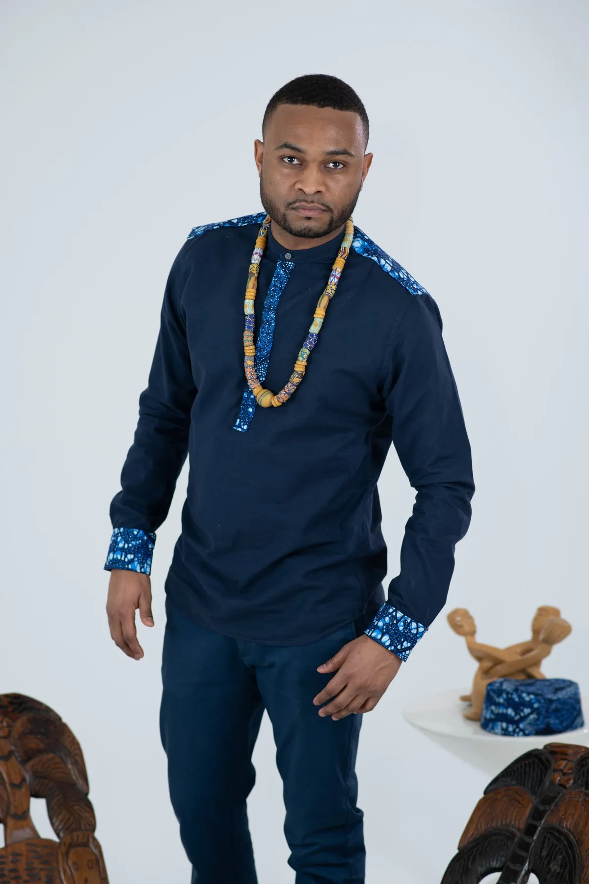 A man in a navy shirt with blue print trim and a beaded necklace stands in the studio
