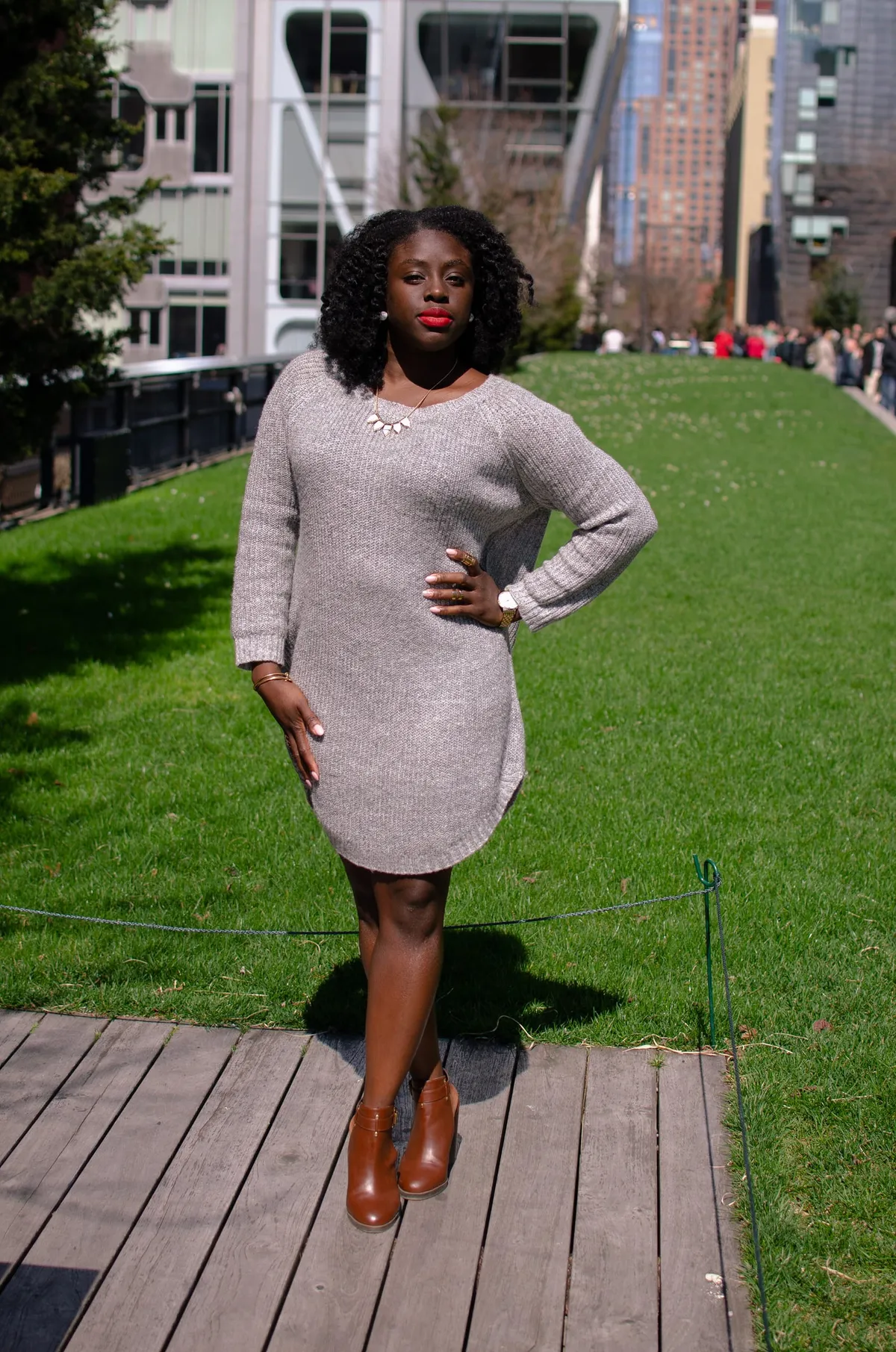 A woman in a grey sweater dress poses on a boardwalk beside a lawn with city buildings behind