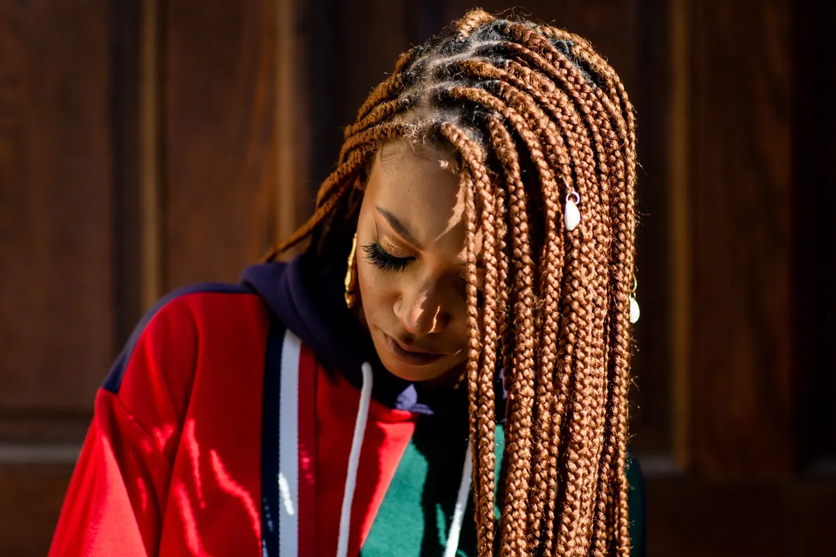 A woman looks down, her long braids falling across a red and green hoodie, in soft side light