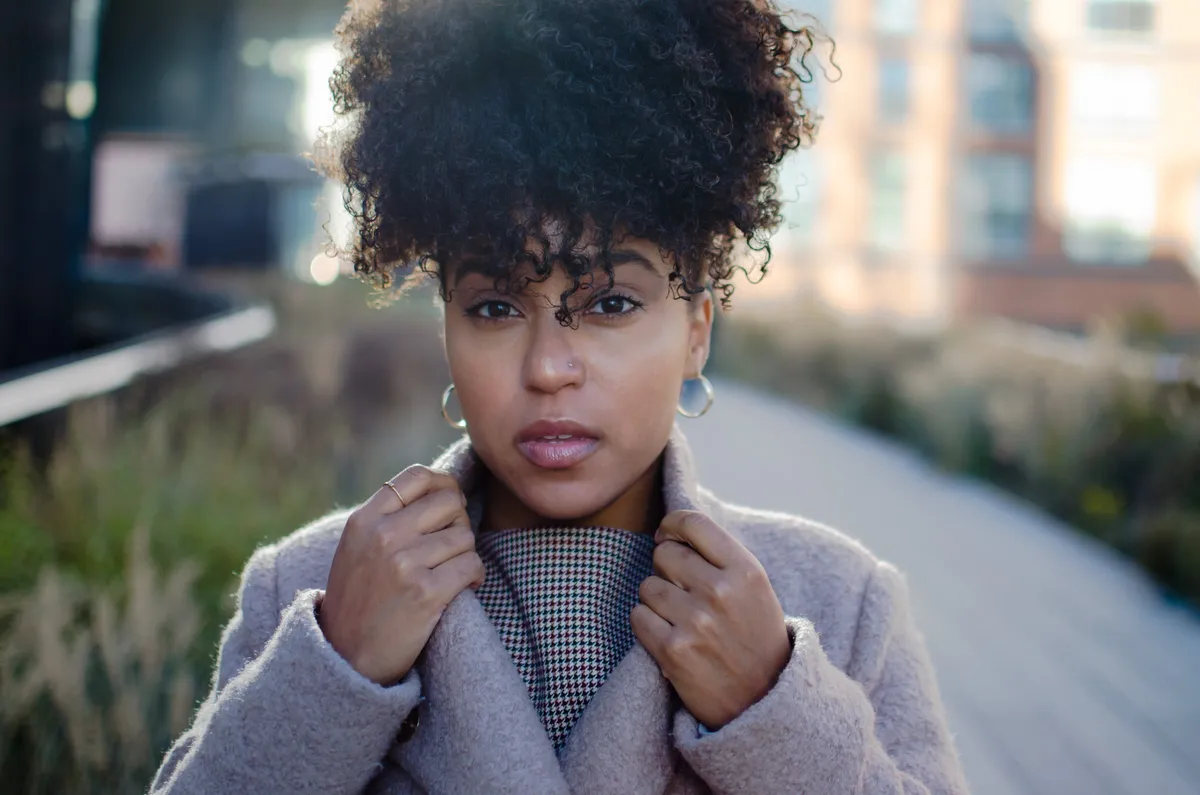 A woman holds the collar of her blush coat on the High Line, gardens blurred behind her