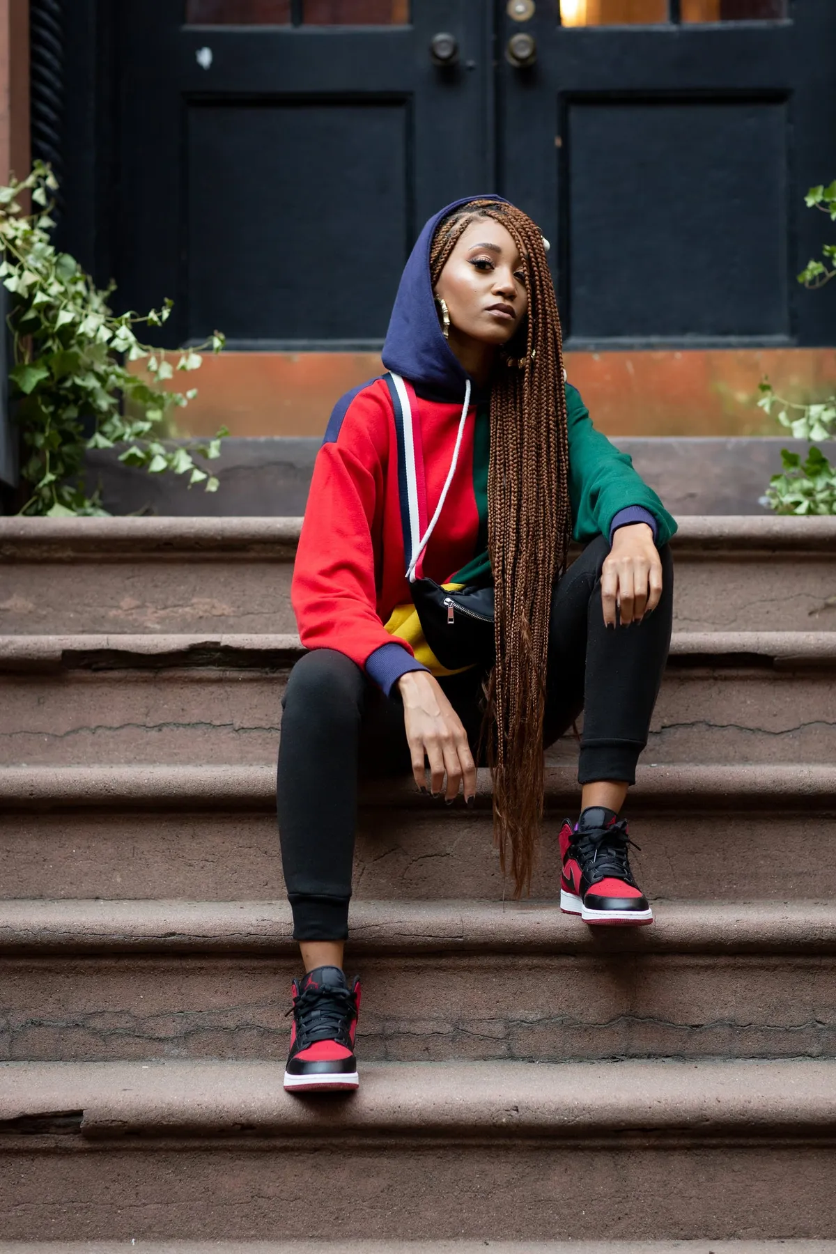 A woman in a hooded color-block sweatshirt sits on brownstone steps beneath a dark green door