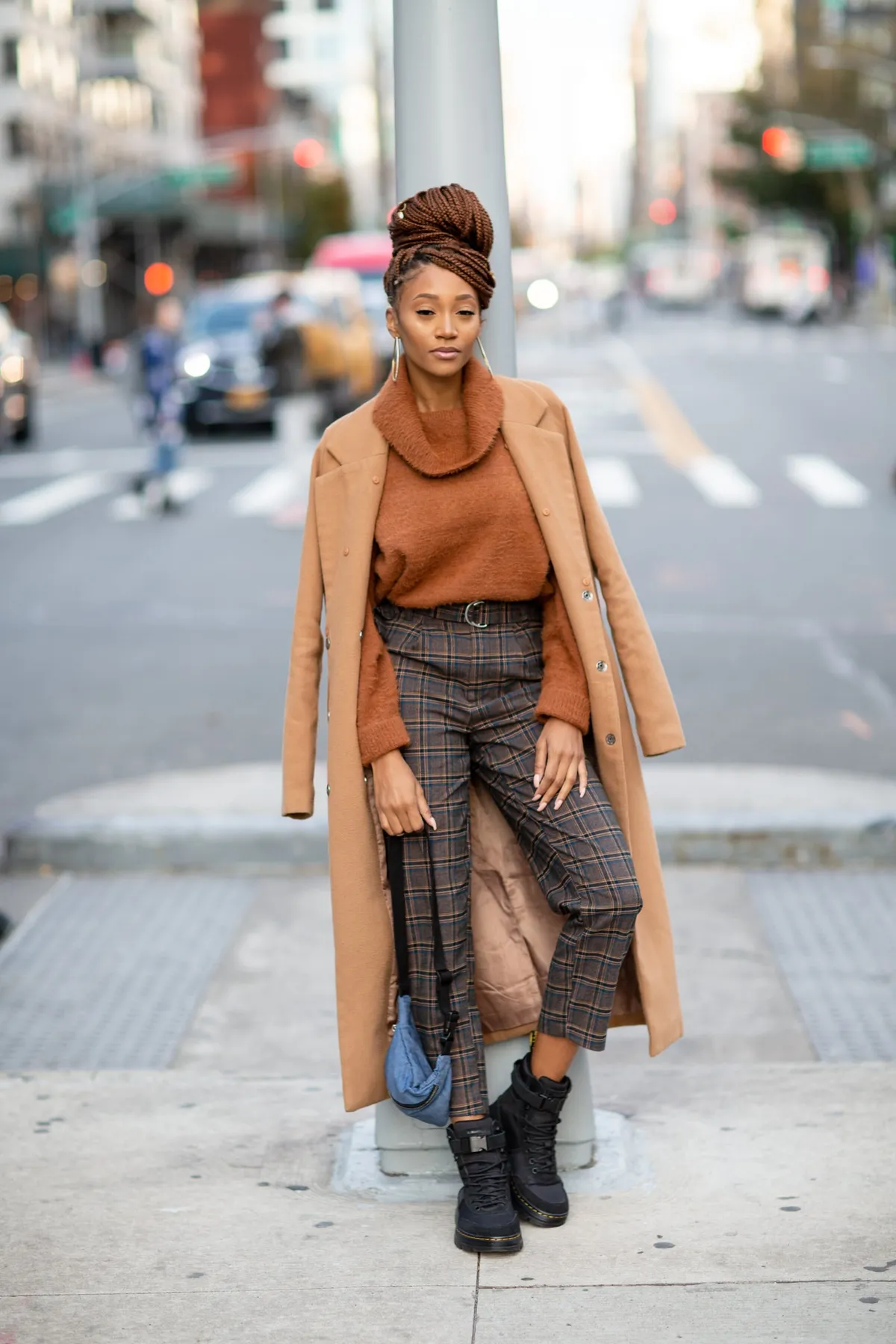 A woman in a camel coat and plaid trousers poses on a city street corner