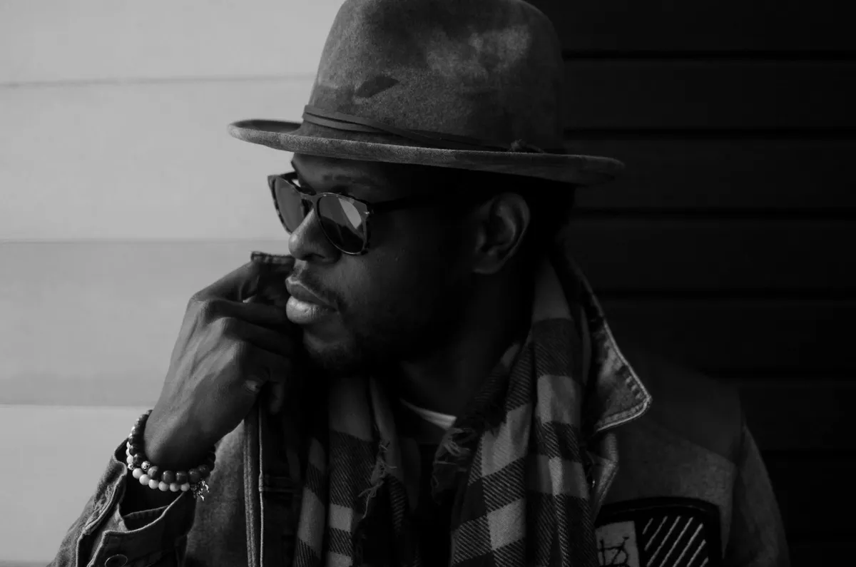 Black-and-white portrait of a man in a fedora and sunglasses with his hand to his face
