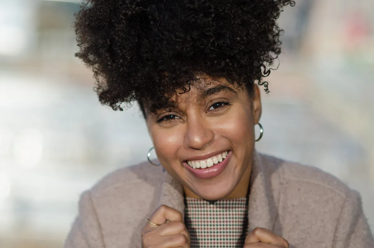 A woman with a natural afro smiles broadly, holding her coat collar