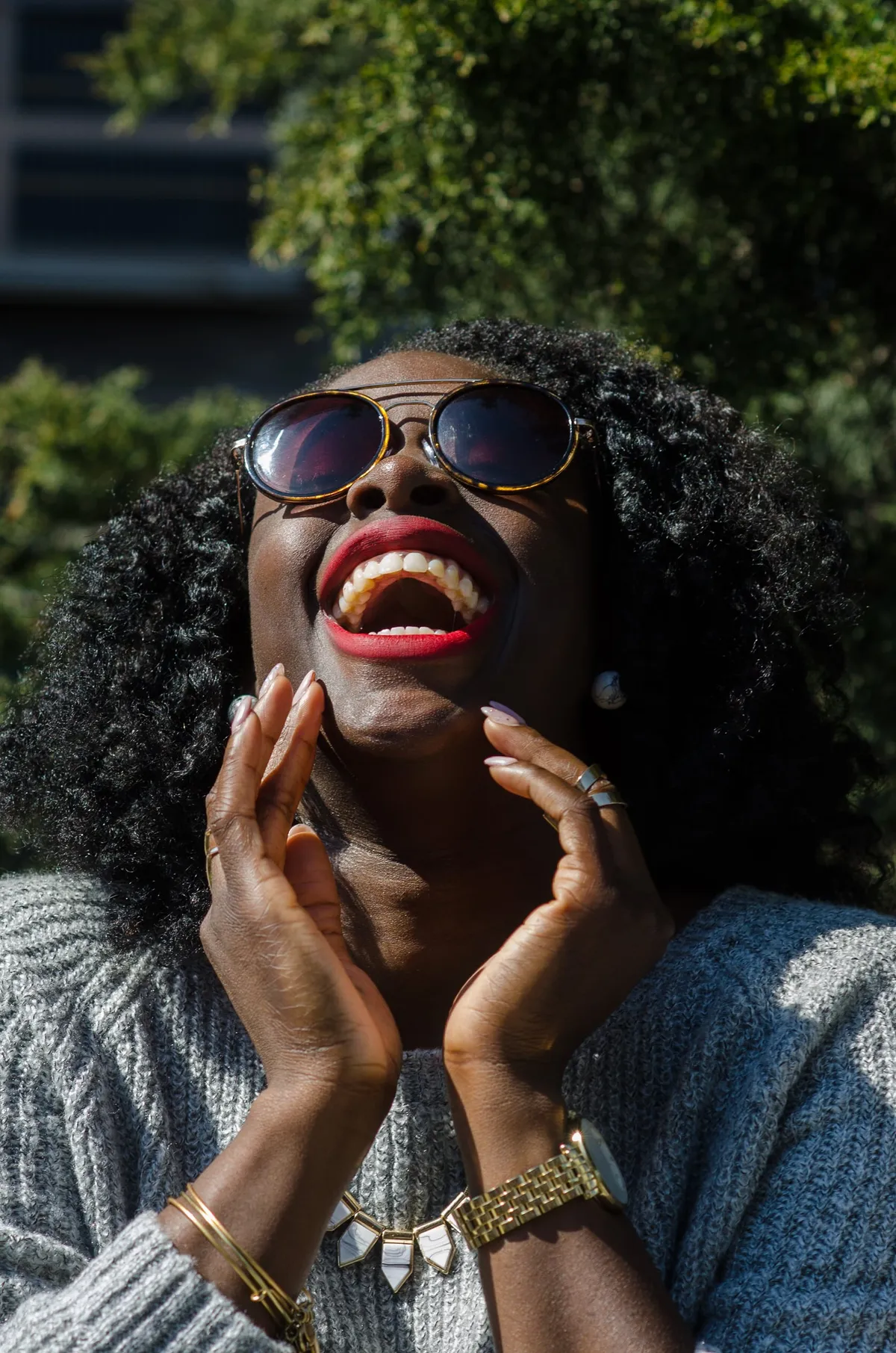 A woman in sunglasses laughs with her head tipped back and her hands at her face