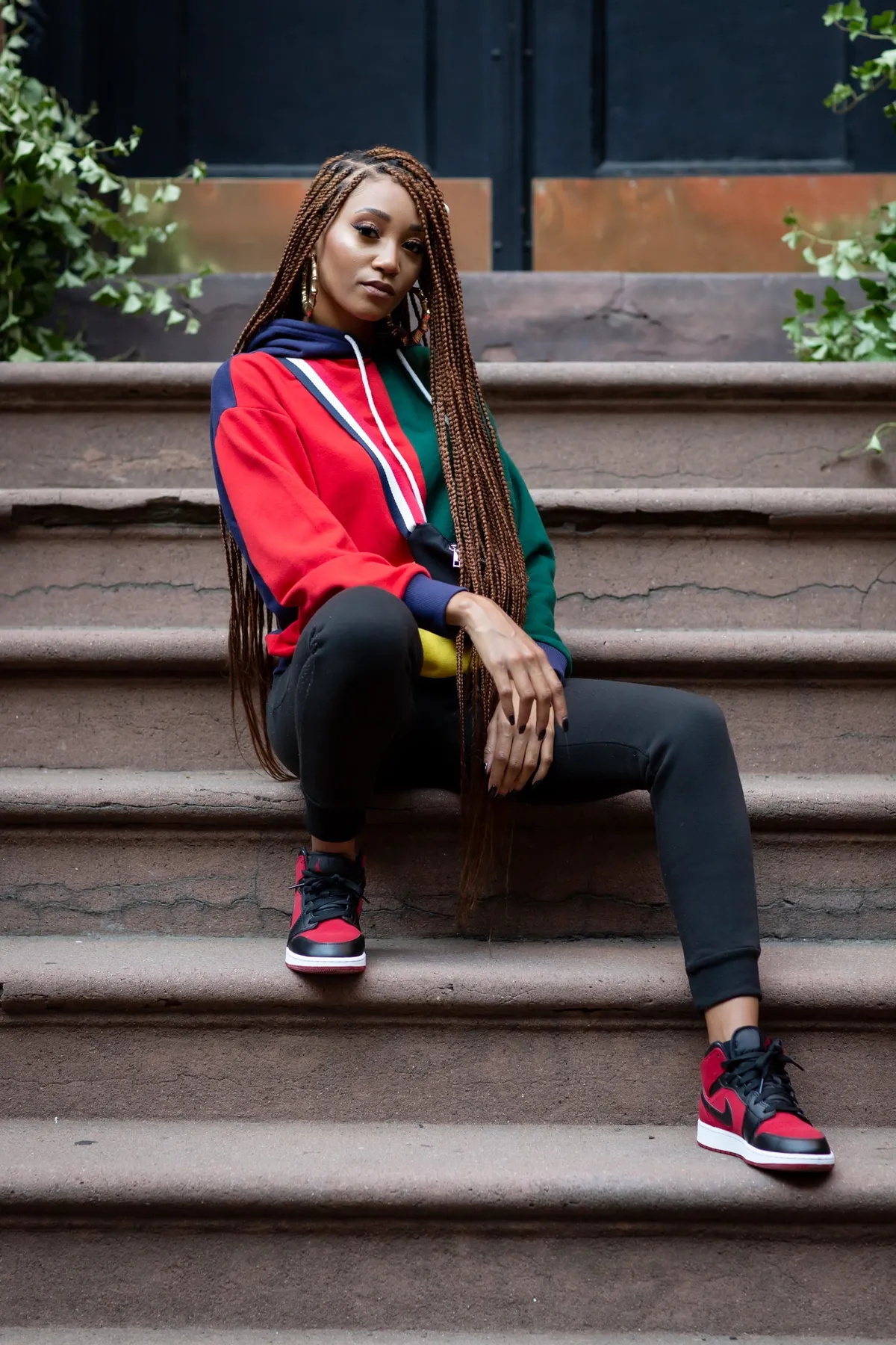A woman leans back on brownstone steps, one leg extended, in a red hoodie and red-and-black sneakers