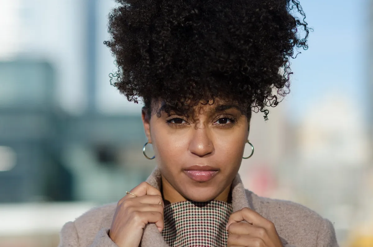 A woman with curly hair and hoop earrings looks into the camera with the city skyline softly blurred